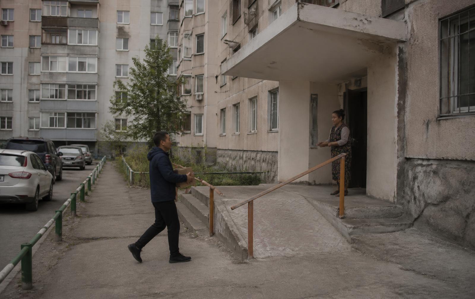 A delivery worker carries a cardboard parcel towards an apartment entrance with a ramp in a residential neighbourhood of Ulaanbaatar.